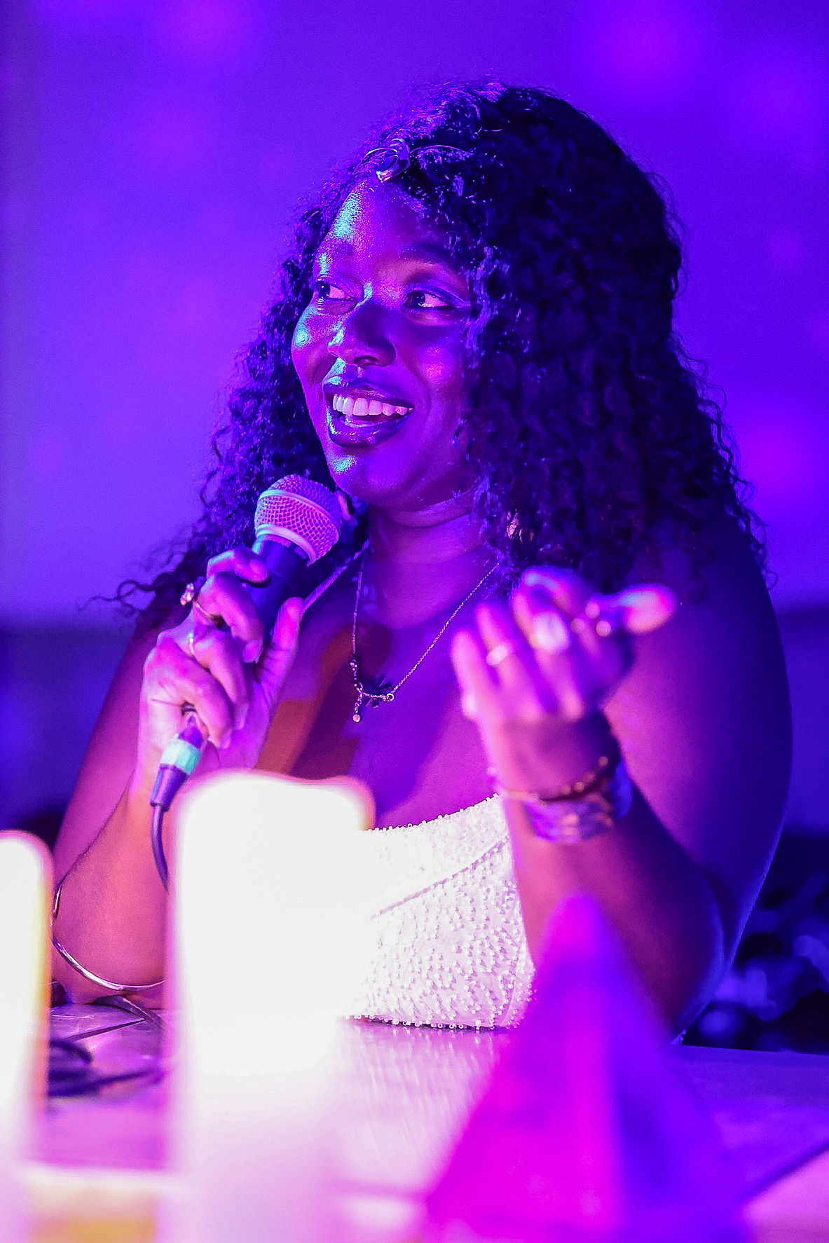 Woman with curly hair smiling, holding a microphone, in a brightly lit setting with purple lighting.