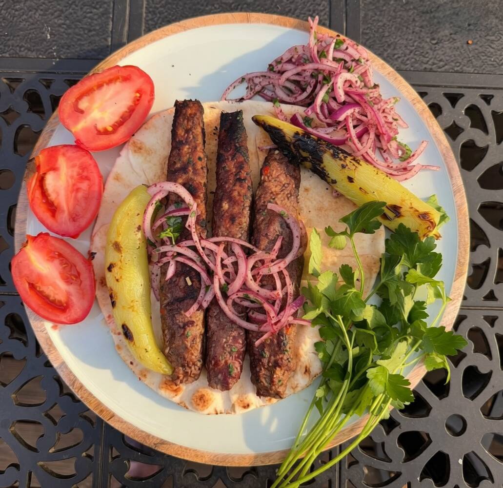 Plate with grilled meat, sliced tomatoes, onion, banana peppers, and cilantro on a decorative black surface.