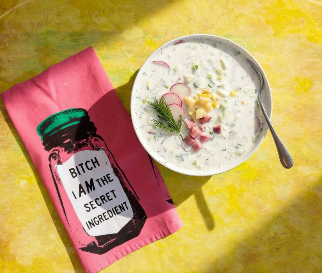 A bowl of creamy soup with herbs and toppings, placed on a yellow surface next to a pink cloth with a jar illustration and text.