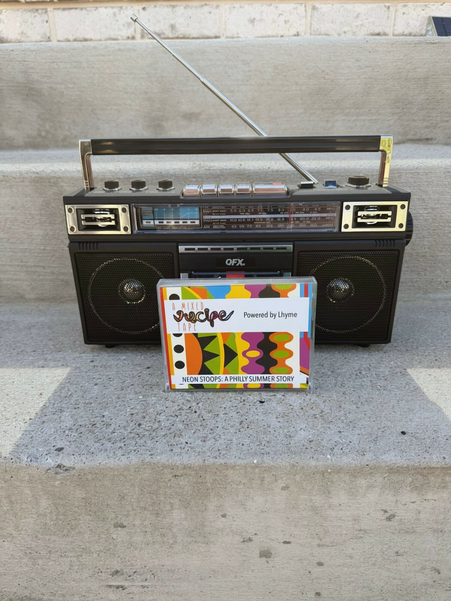 Portable radio and cassette player with a colorful card in front, placed on concrete surface outdoors.