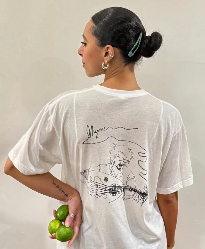 Woman with dark hair in bun, wearing a white T-shirt with line art, holding limes, facing left.