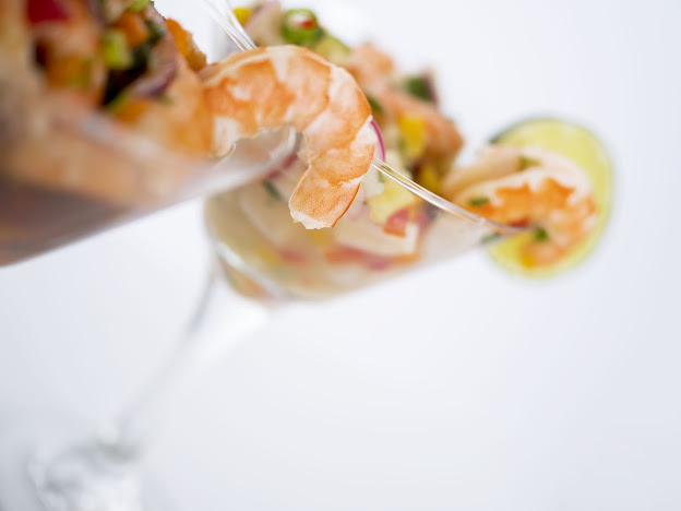 A close-up of a shrimp being placed into a glass filled with seafood salad, with a lemon wedge in the background.