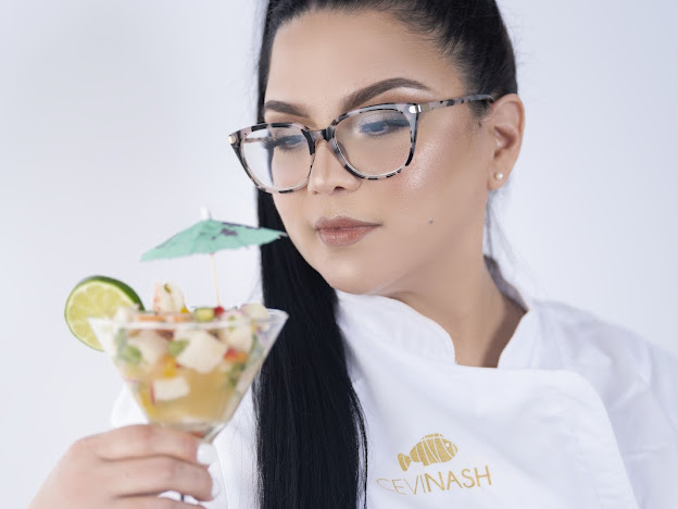 Woman with glasses holding a cocktail with a small umbrella and lime slice, wearing a white shirt with a logo.