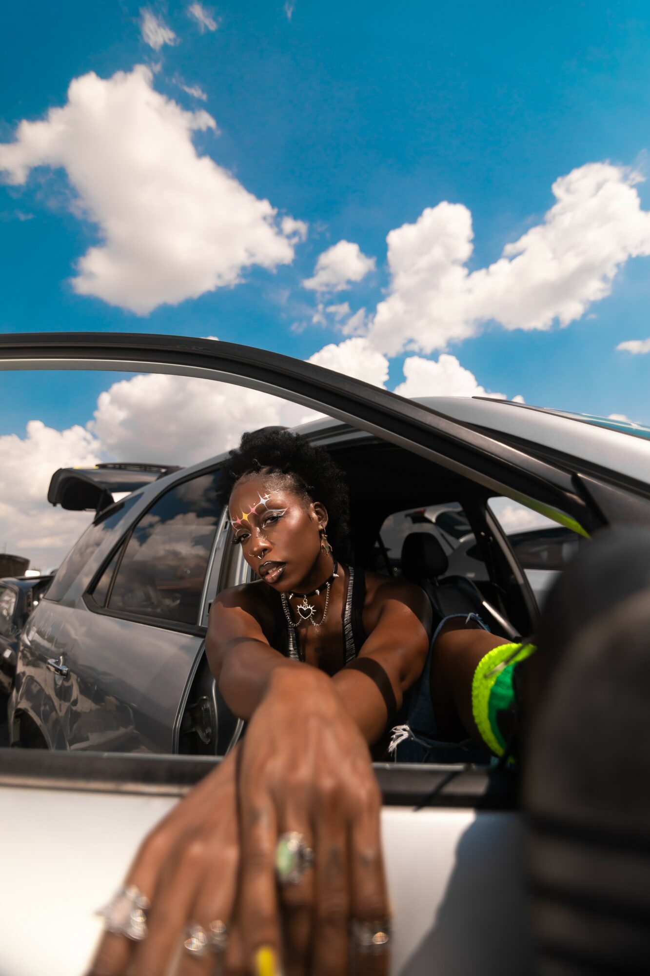 Person with jewelry reaching out of a car window, with a blue sky and clouds in the background.