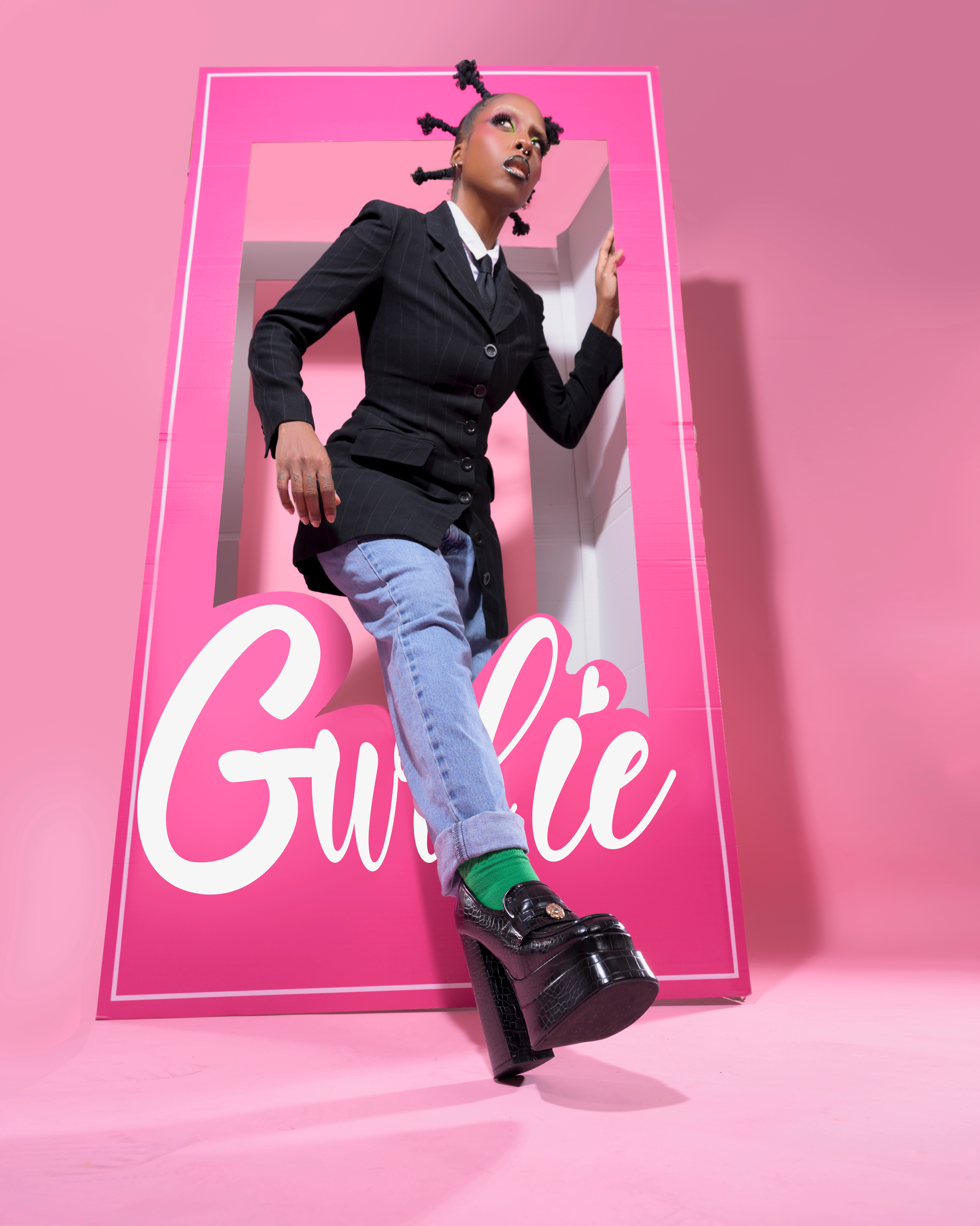Person with styled hair in a black blazer and jeans stands in a pink box with 'Gurlie' written on it, pink background.