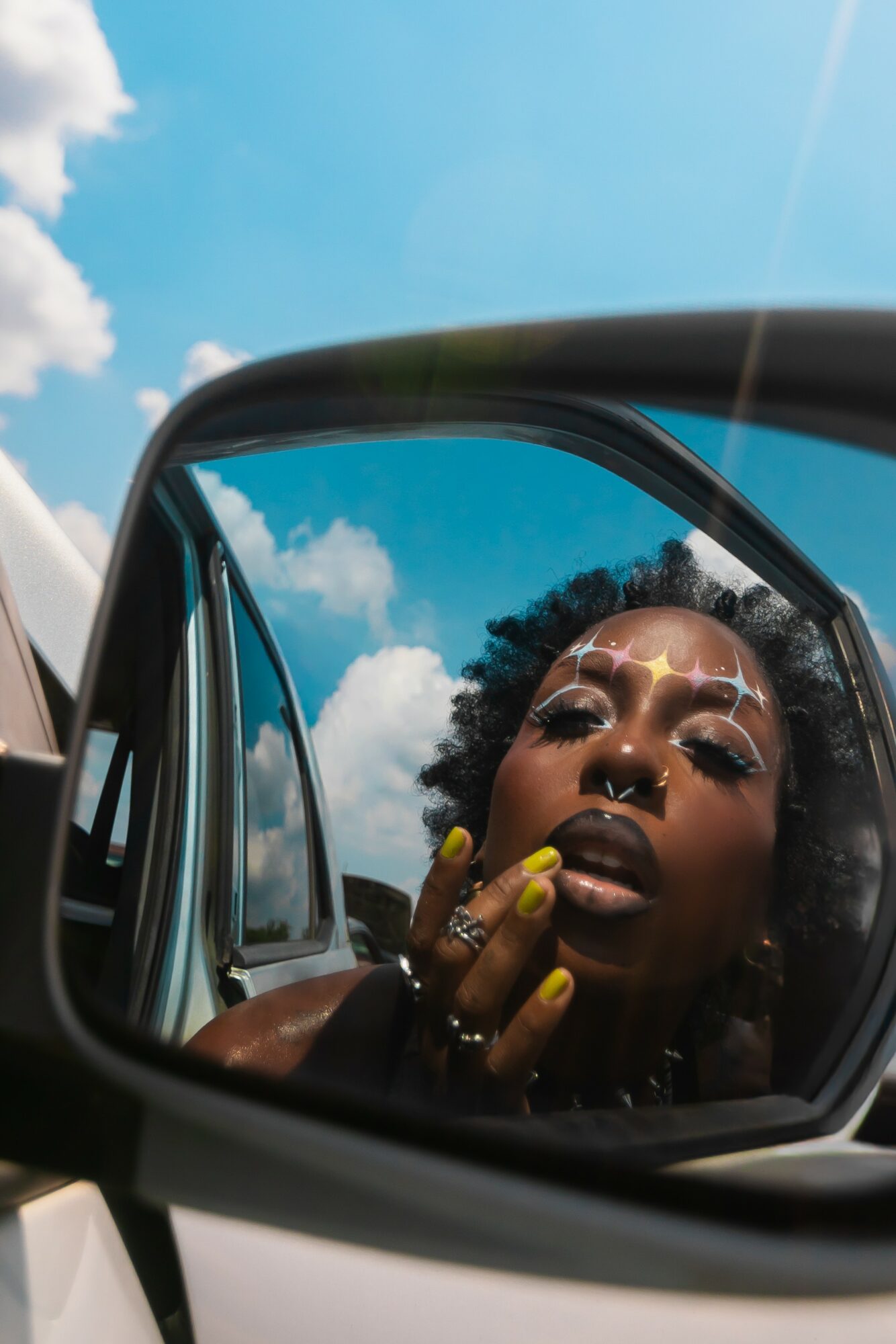 Person with makeup and yellow nail polish looking into car side mirror, reflection shows blue sky and clouds