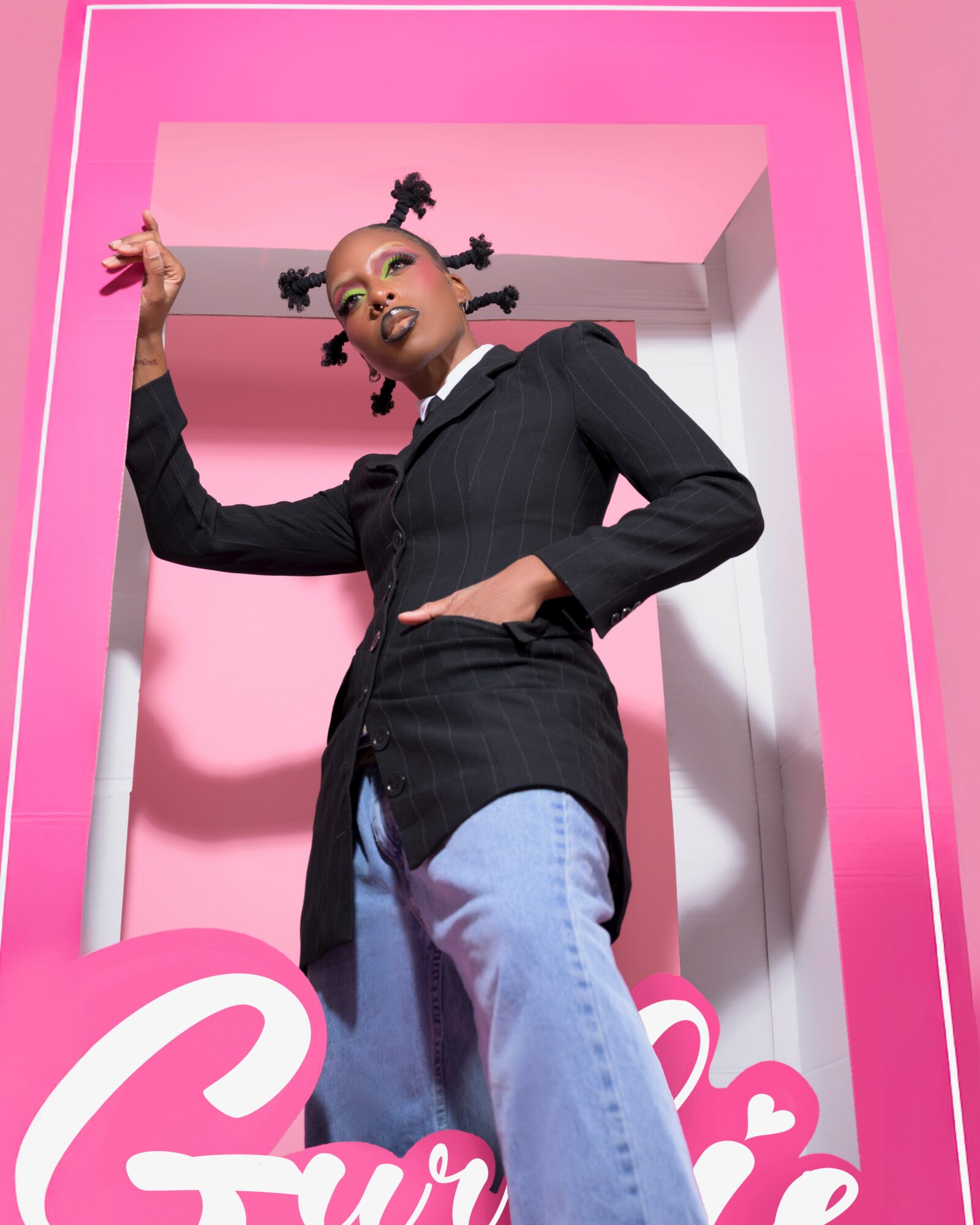 Young woman with styled hair in a black blazer and jeans, standing in a pink geometric frame, looking at the camera.