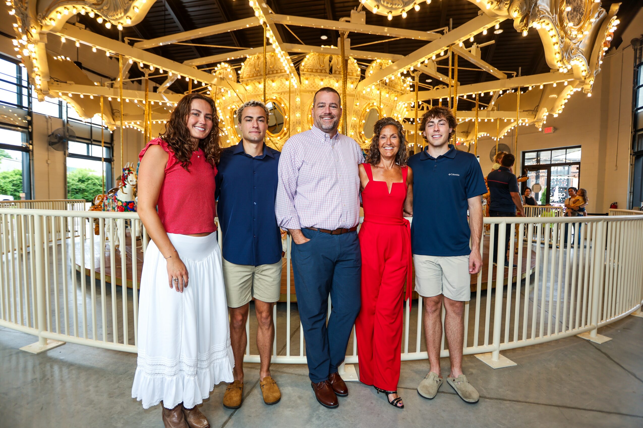 Group of five people standing in front of a carousel with lights, smiling, indoors, casual and semi-formal attire.