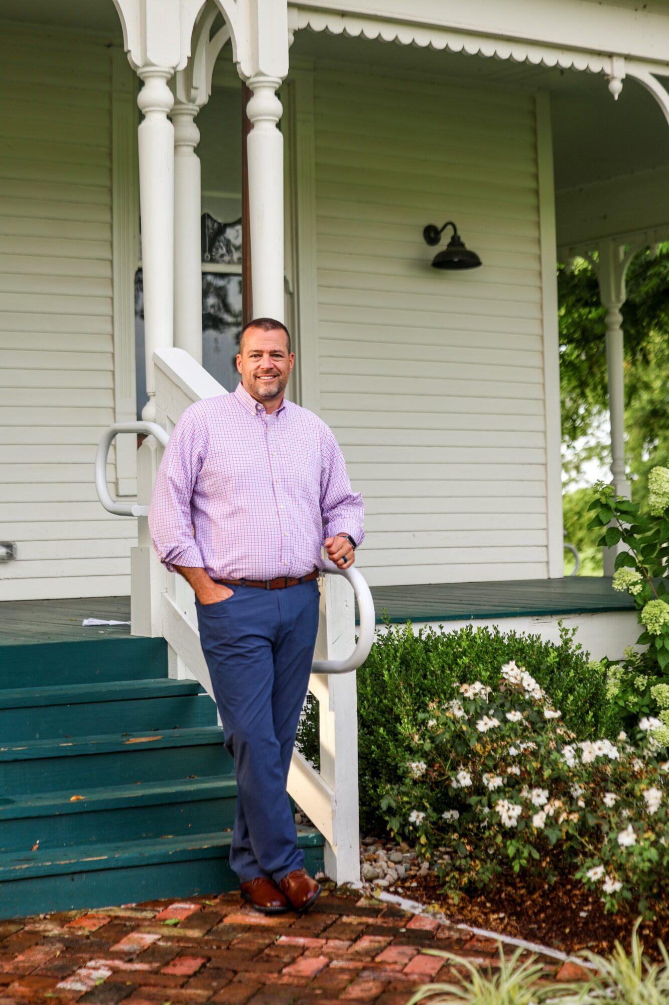 Man in purple shirt and blue pants standing on porch steps with garden and house in background.