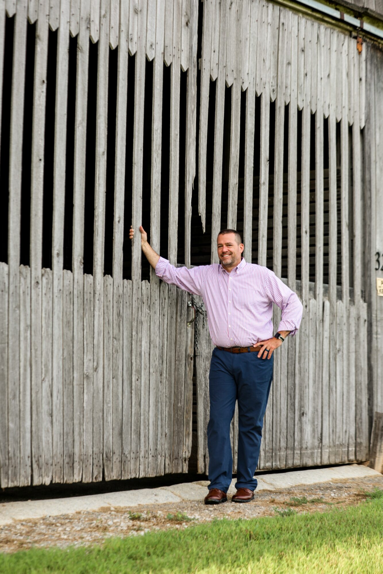 Man in pink shirt and blue jeans standing next to a tall metal fence, smiling, with one hand on the fence.