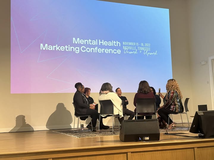 Six people seated on stage in front of a large screen displaying 'Mental Health Marketing Conference' and event details.