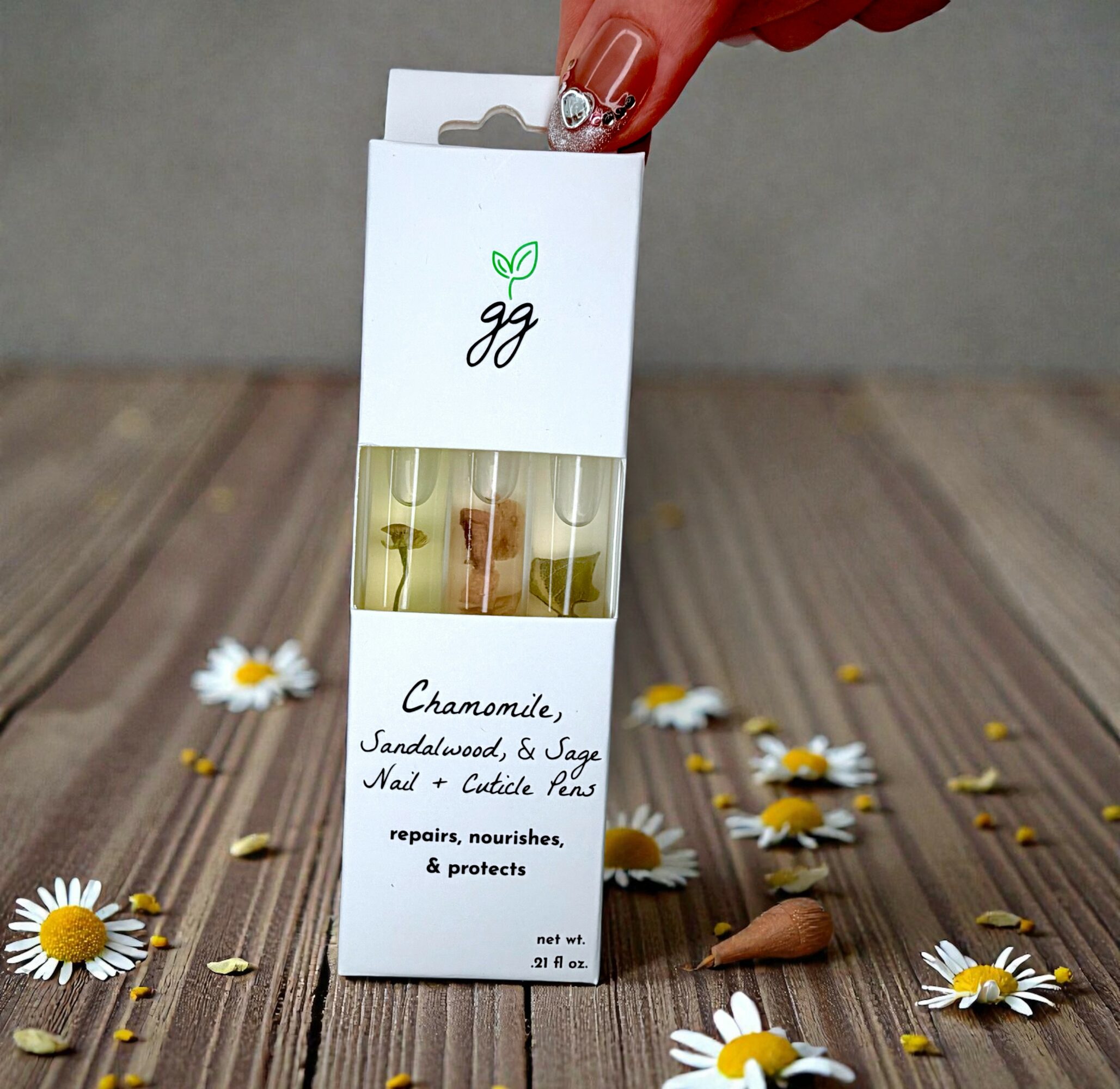Hand holding a small white box with a clear window showing essential oils, surrounded by daisies on a wooden surface.