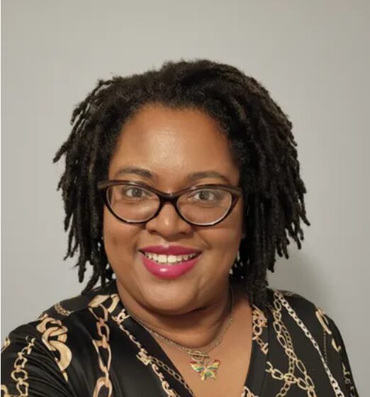 Portrait of a woman with glasses, smiling, wearing a black and gold patterned top, and a necklace with a butterfly pendant.