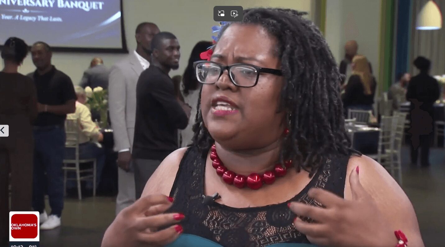 Woman with glasses and curly hair speaking at an event, wearing a black lace top and red necklace.