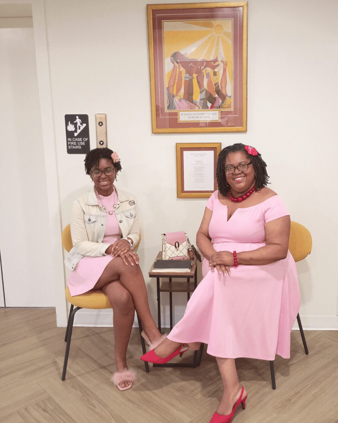 Two women in pink dresses and floral headbands sitting on chairs in a room with framed artwork and a small table between them.
