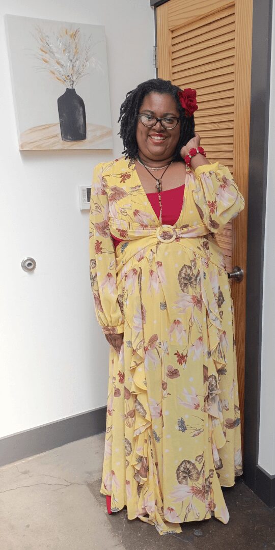 Woman in yellow floral dress with red top, standing indoors, smiling, with artwork and wooden door behind her.