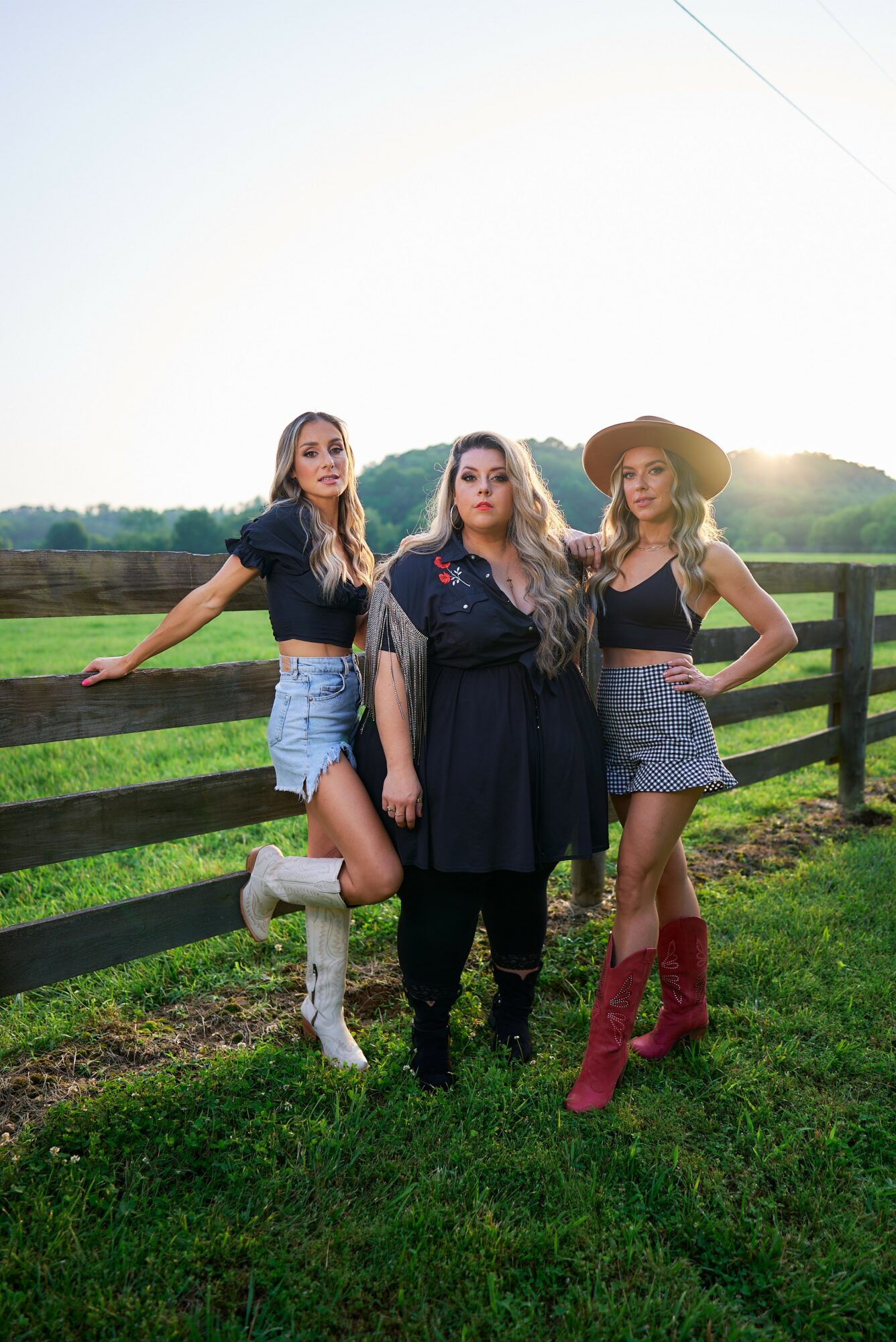 Three women standing outdoors near a wooden fence with a field and trees in the background, sunlight shining behind them.