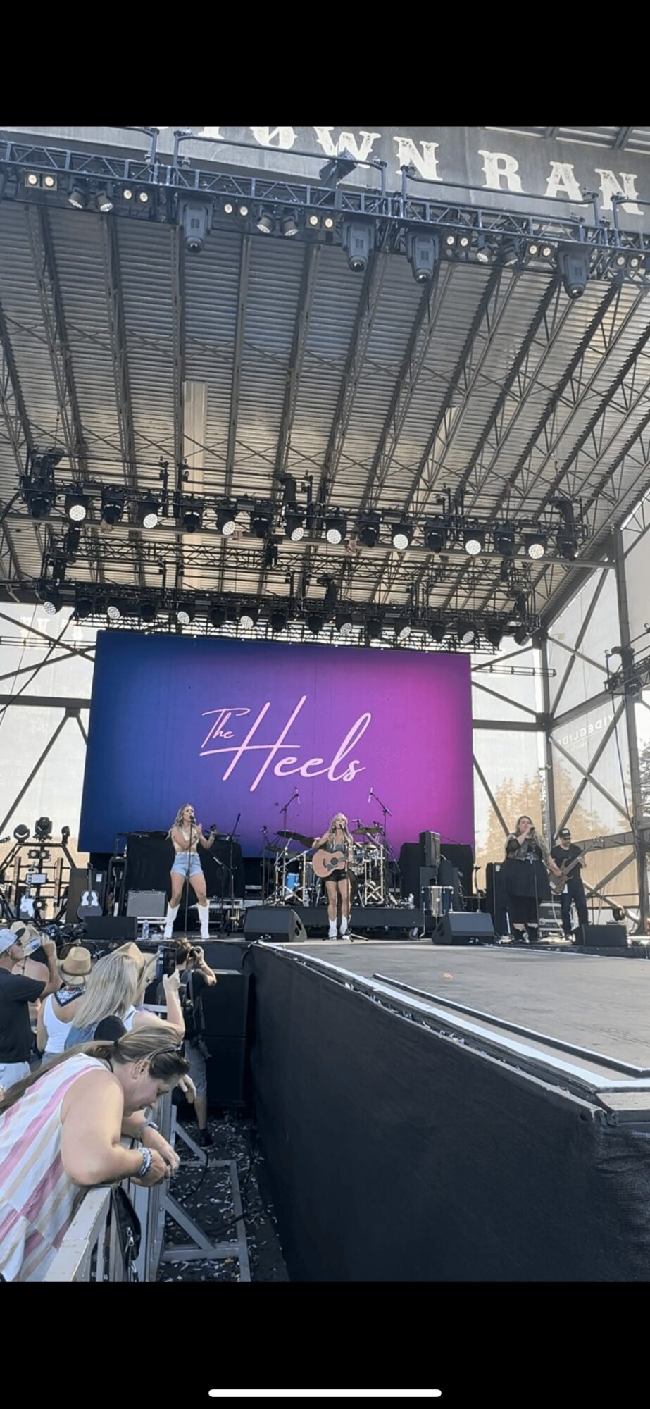 Stage with performers and a large screen displaying 'Hello' in pink, outdoor setting with audience and equipment.
