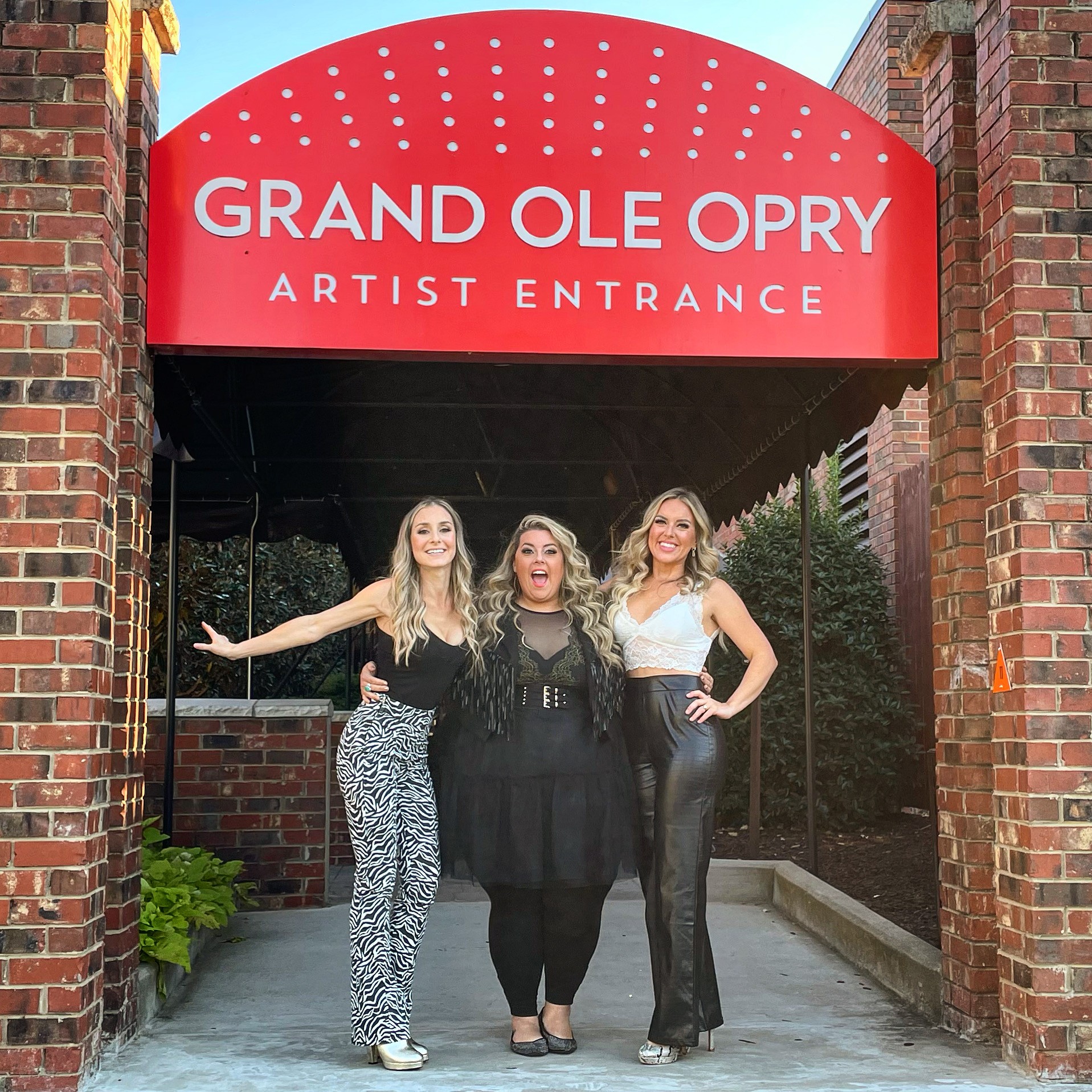 Three women standing under a red sign that reads 'Grand Ole Opry Artist Entrance' outside brick building, smiling.