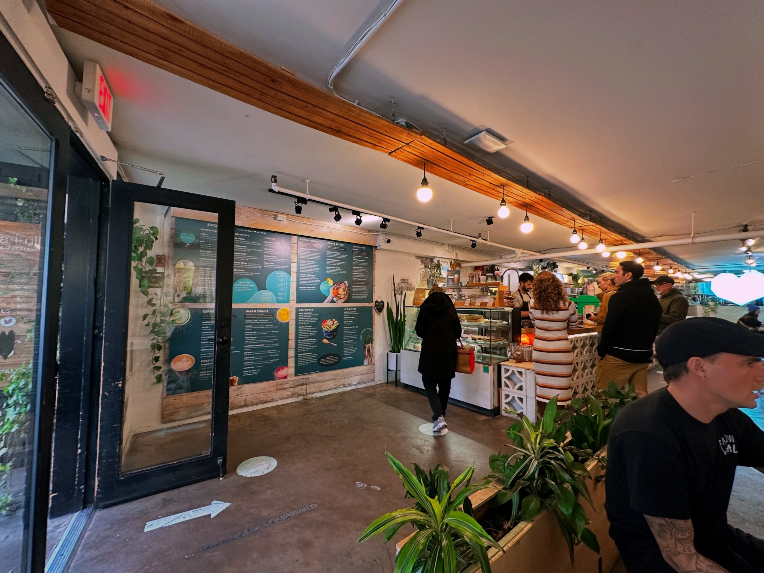 People inside a store or cafe with plants and shelves, some browsing and others working or talking, warm lighting, wooden ceiling.