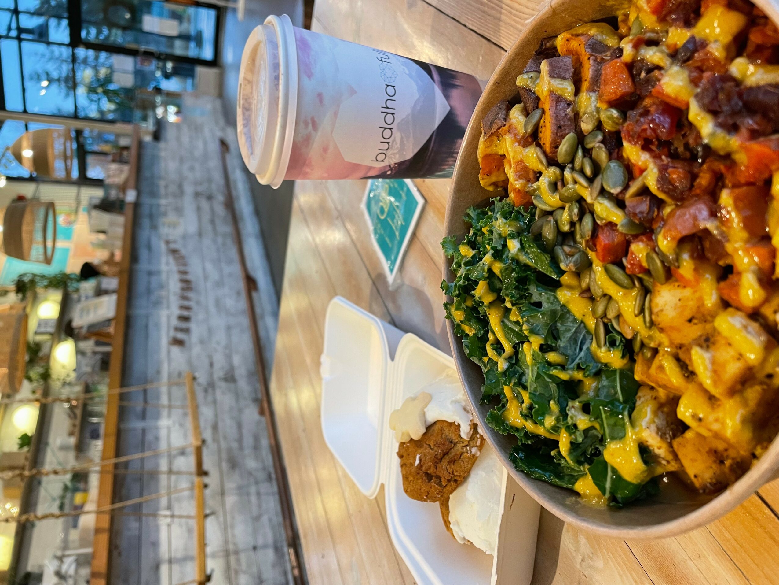 Bowl of pasta with vegetables and kale, a drink, and a small plate with condiments on a wooden table.