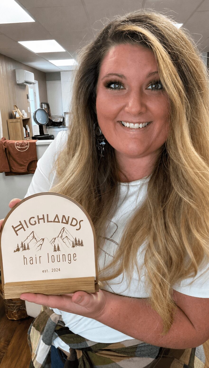 Woman with long, wavy hair holding a sign that reads 'Highlands Hair Lounge' in a salon setting.