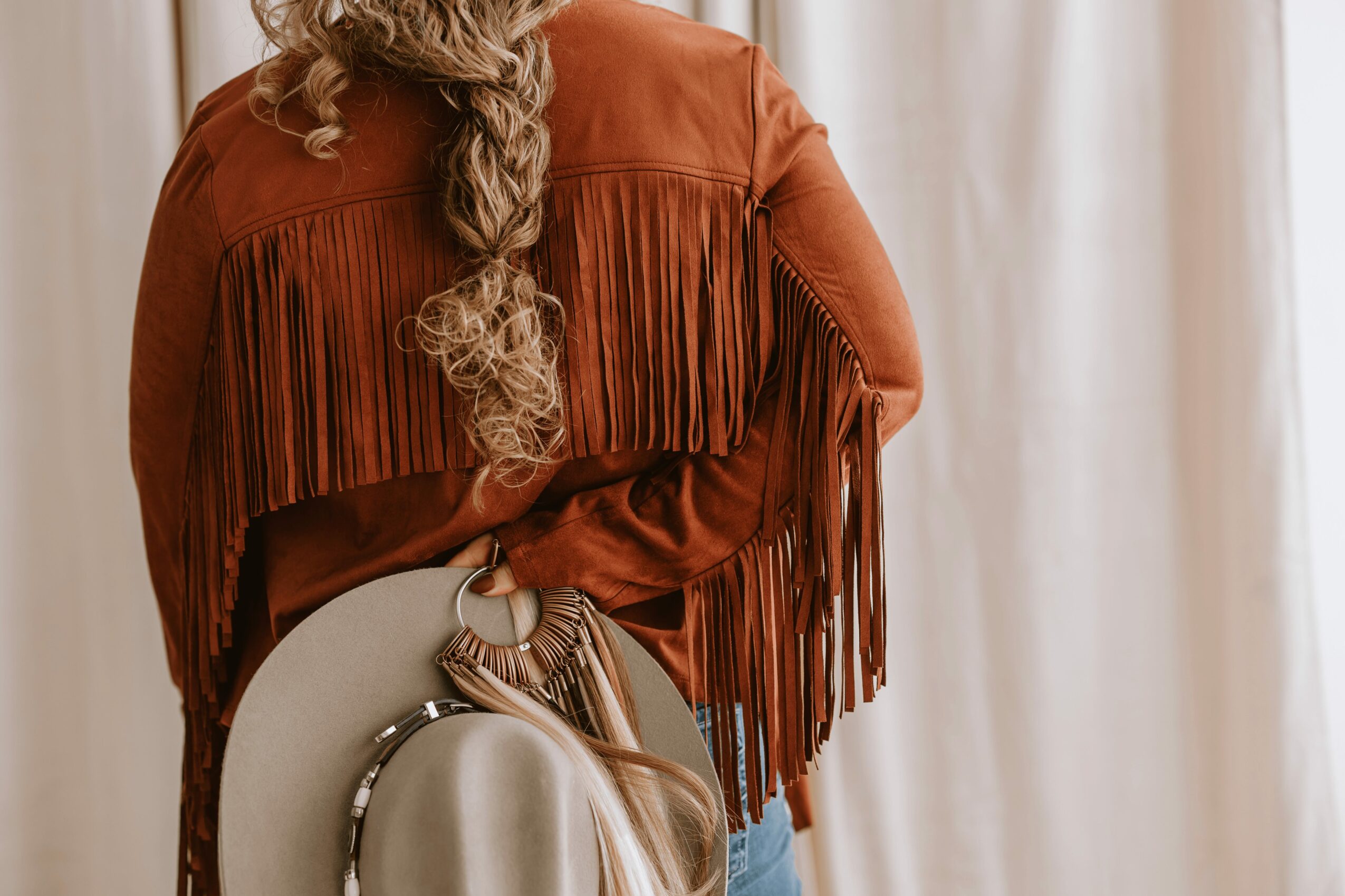 Person with long curly hair wearing a fringed brown jacket, standing near a curtain, holding a round mirror with a beige strap.