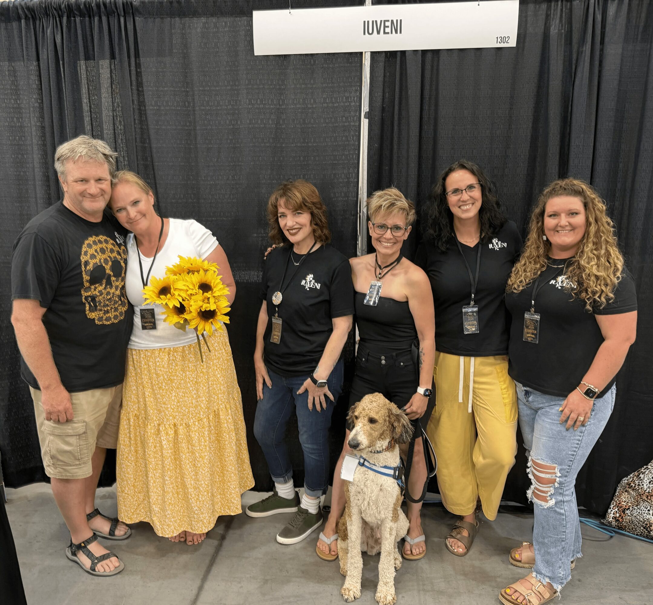Six people and a dog stand in front of a black curtain, with a sign reading 'IUVENI' above them.