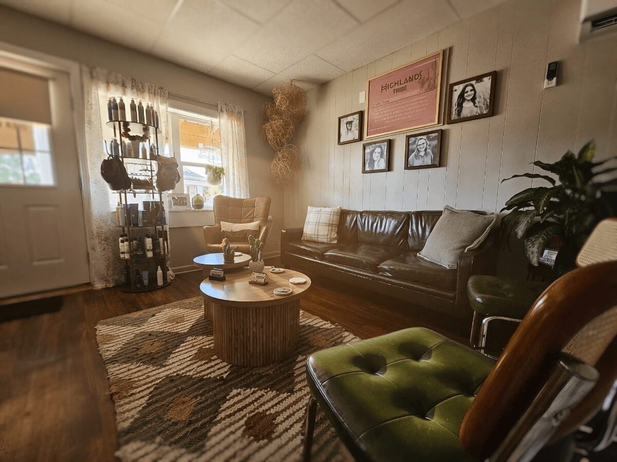 Living room with a black sofa, green armchair, round coffee table, and wall art, sunlight through windows, and houseplants.