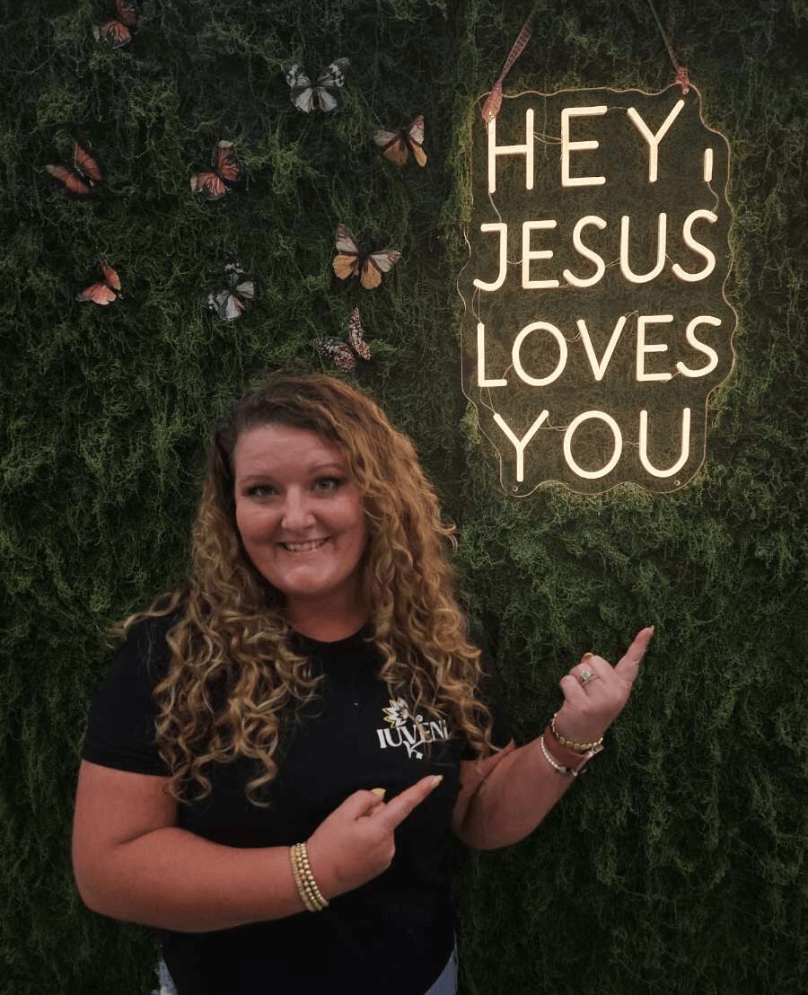 Woman with curly hair smiling, pointing at a sign that says 'Hey Jesus Loves You,' against a green leafy background.