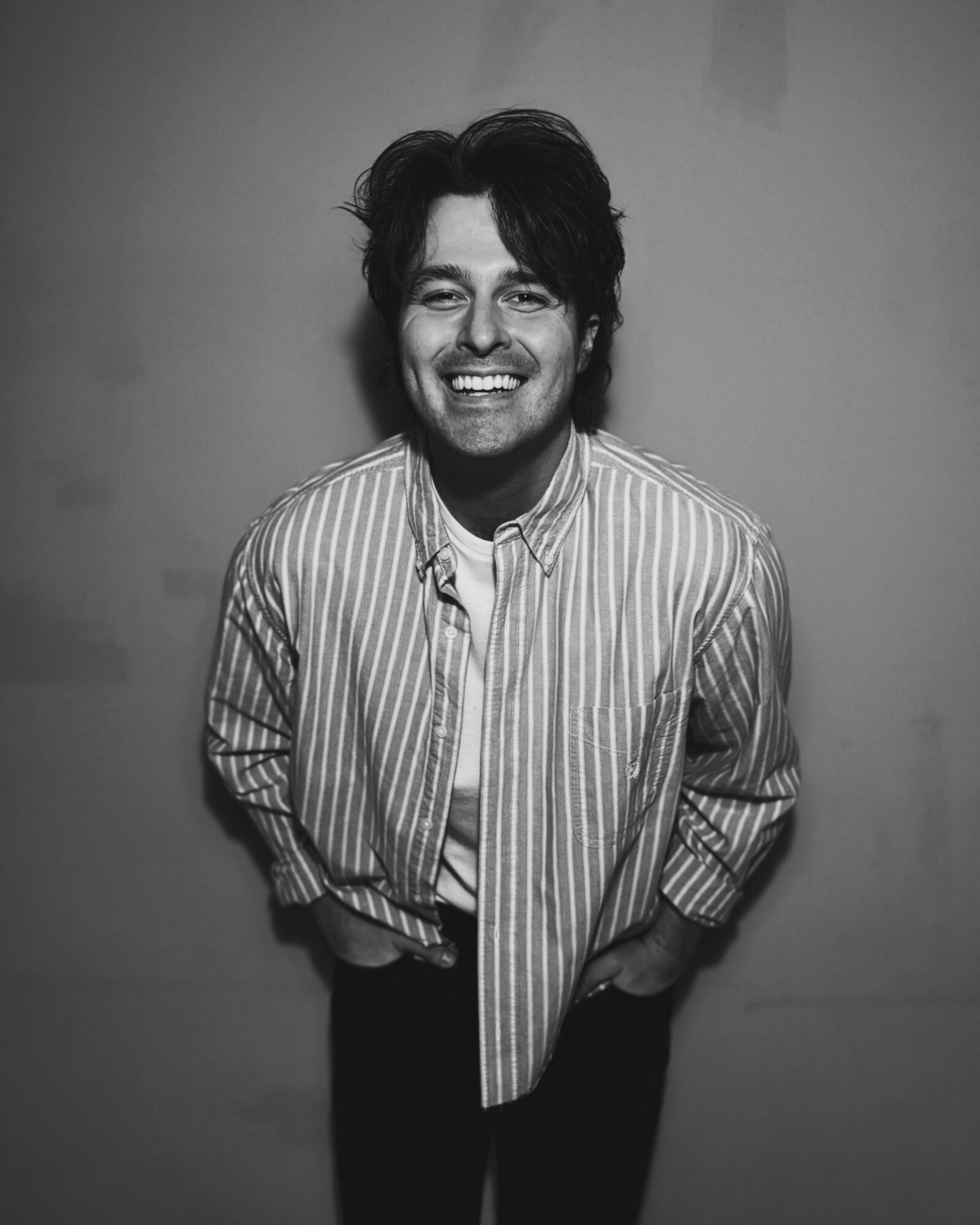 Smiling person with dark, wavy hair wearing a striped shirt, standing against a plain background.