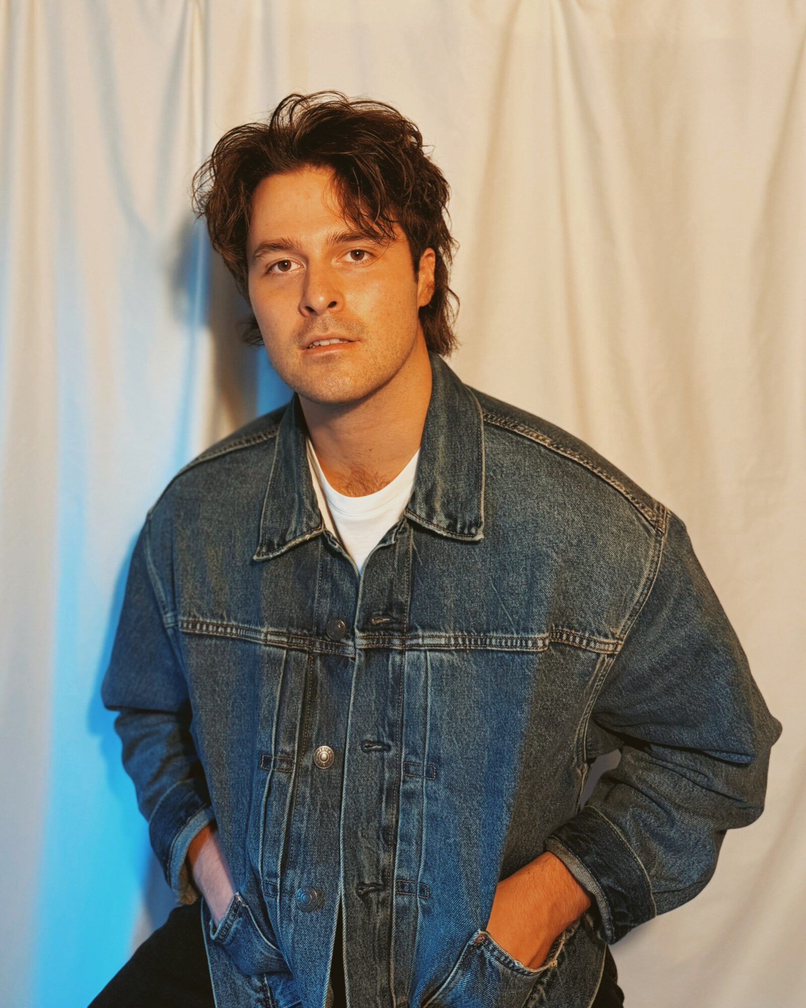Young man with wavy hair wearing a denim jacket and white shirt, standing in front of a cream-colored curtain.