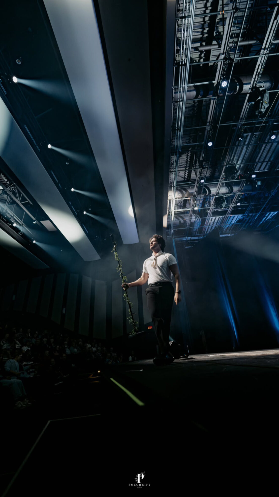 Person standing on stage with overhead lighting and ceiling structure visible, audience in darkness, stage illuminated.