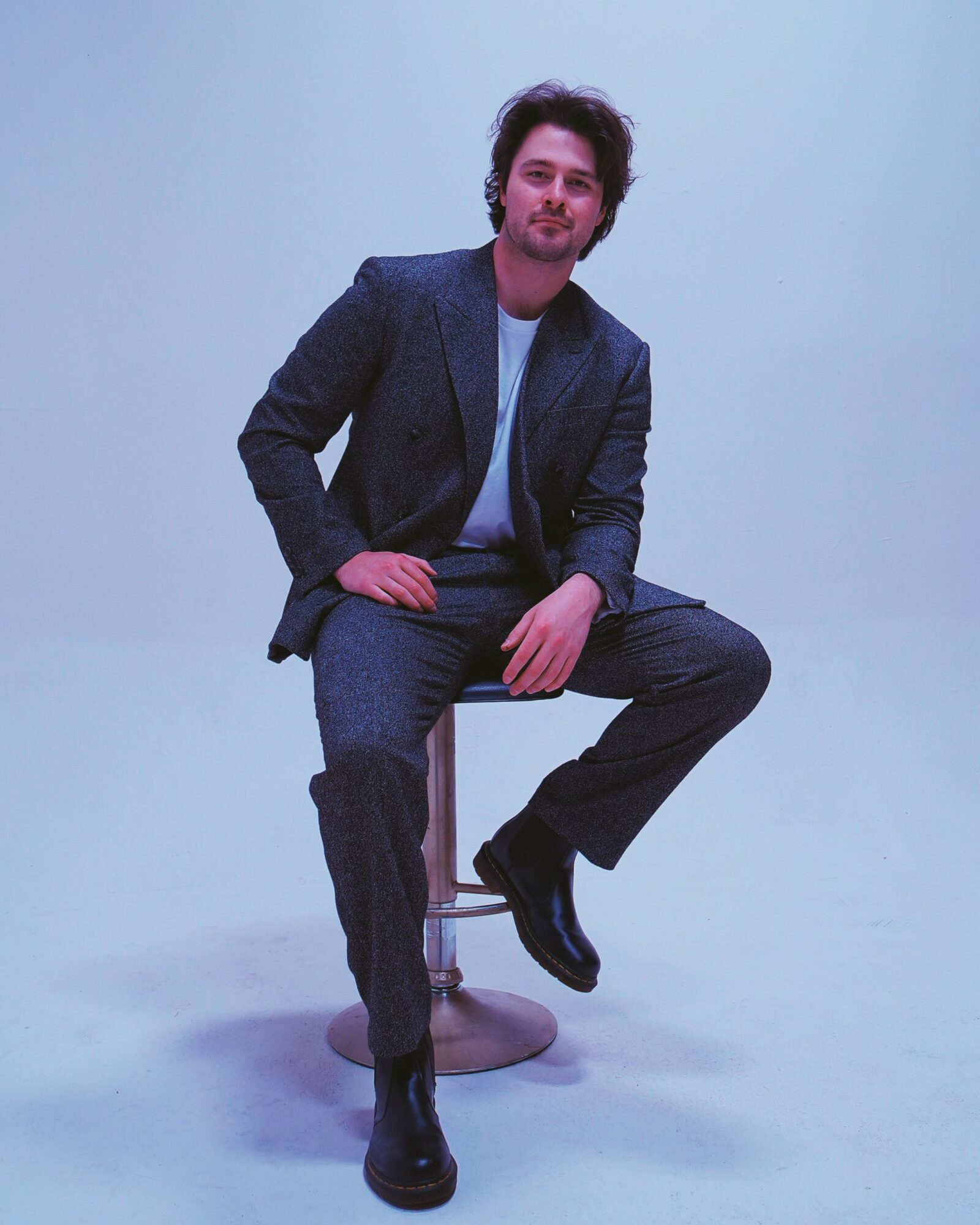 Man with dark hair and beard sitting on a stool, wearing a blazer, jeans, and black shoes, against a plain light background.