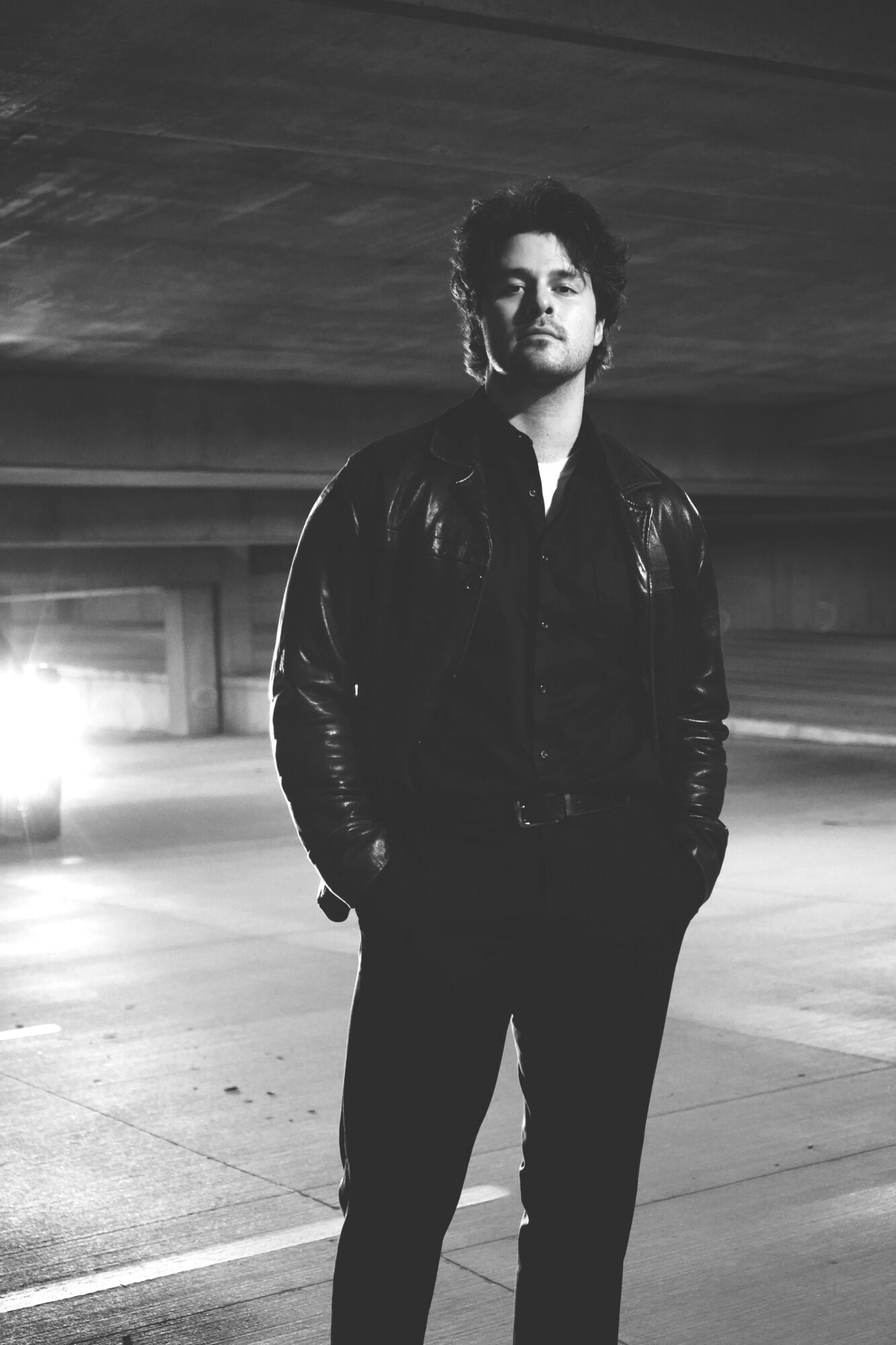 Young man standing in an empty parking garage, wearing a leather jacket and dark pants, with hands in pockets.