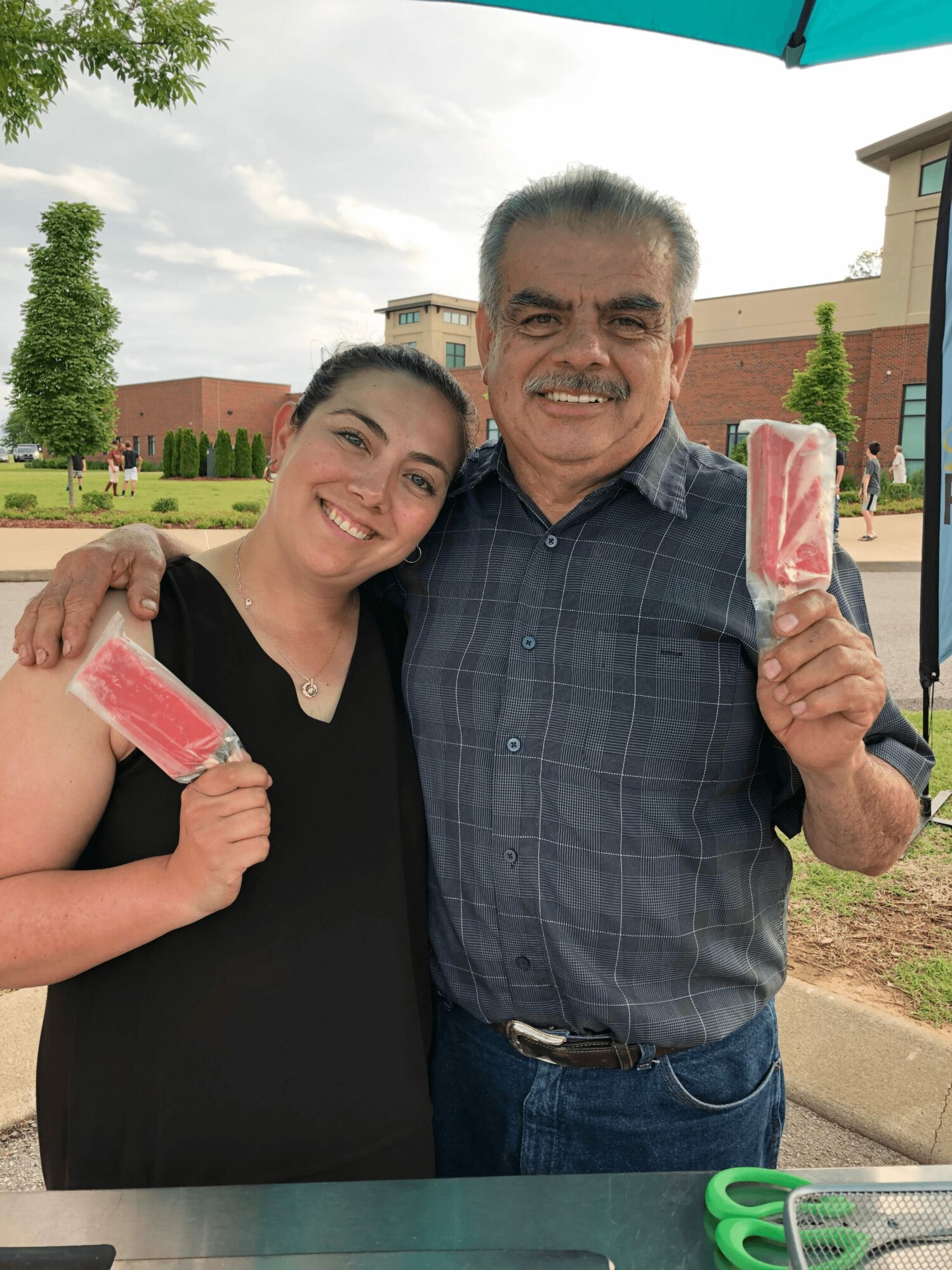 Two people smiling outdoors, holding pink popsicles, with trees and buildings in the background.