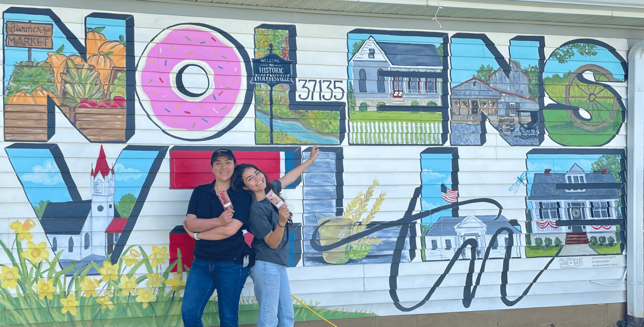 Two women stand in front of a colorful mural with large letters spelling 'MORGAN'S VIEWS' and a house scene, smiling.