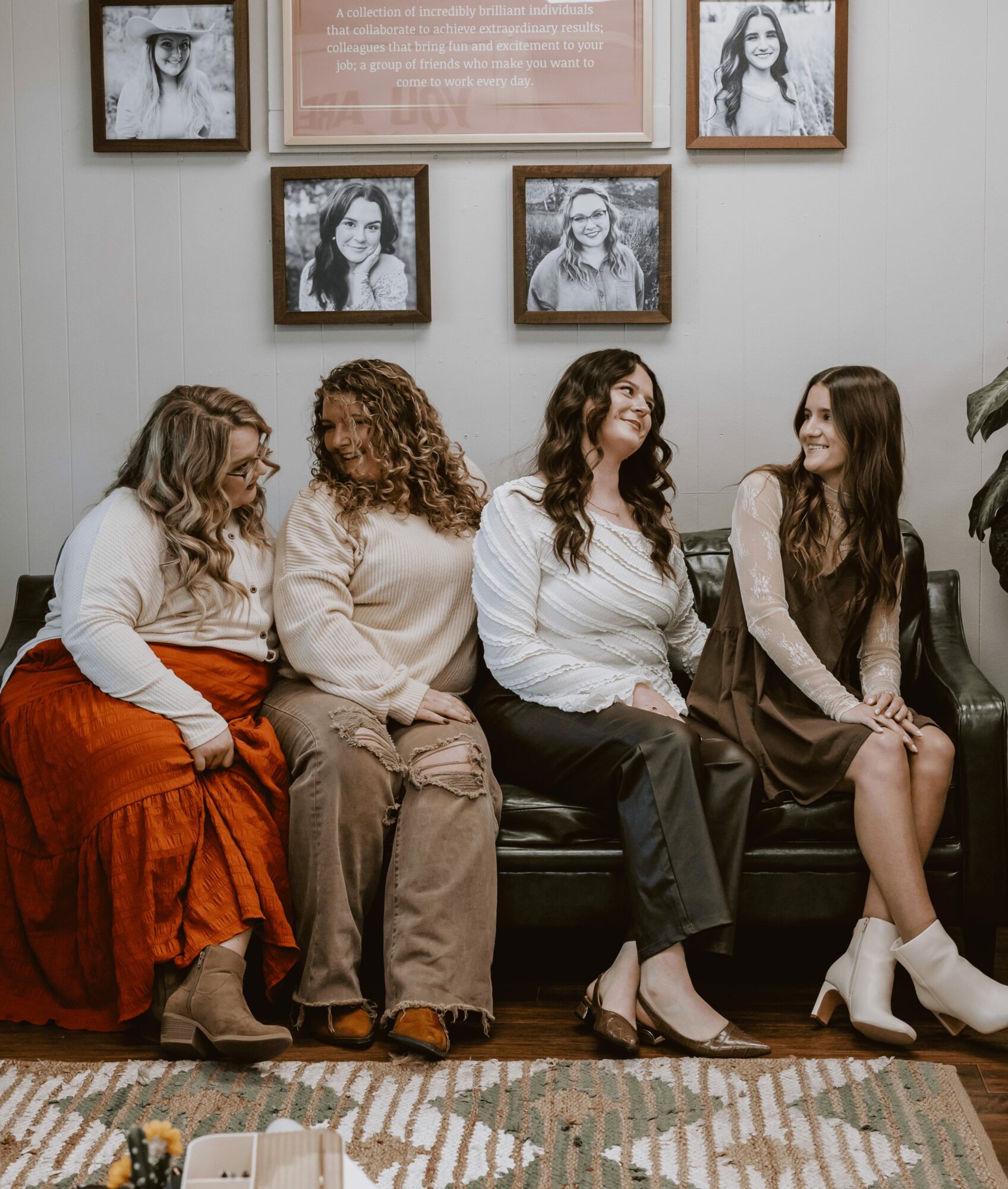 Four women sitting on a black sofa, smiling, in a room with framed photos on the wall, a plant, and a patterned rug.