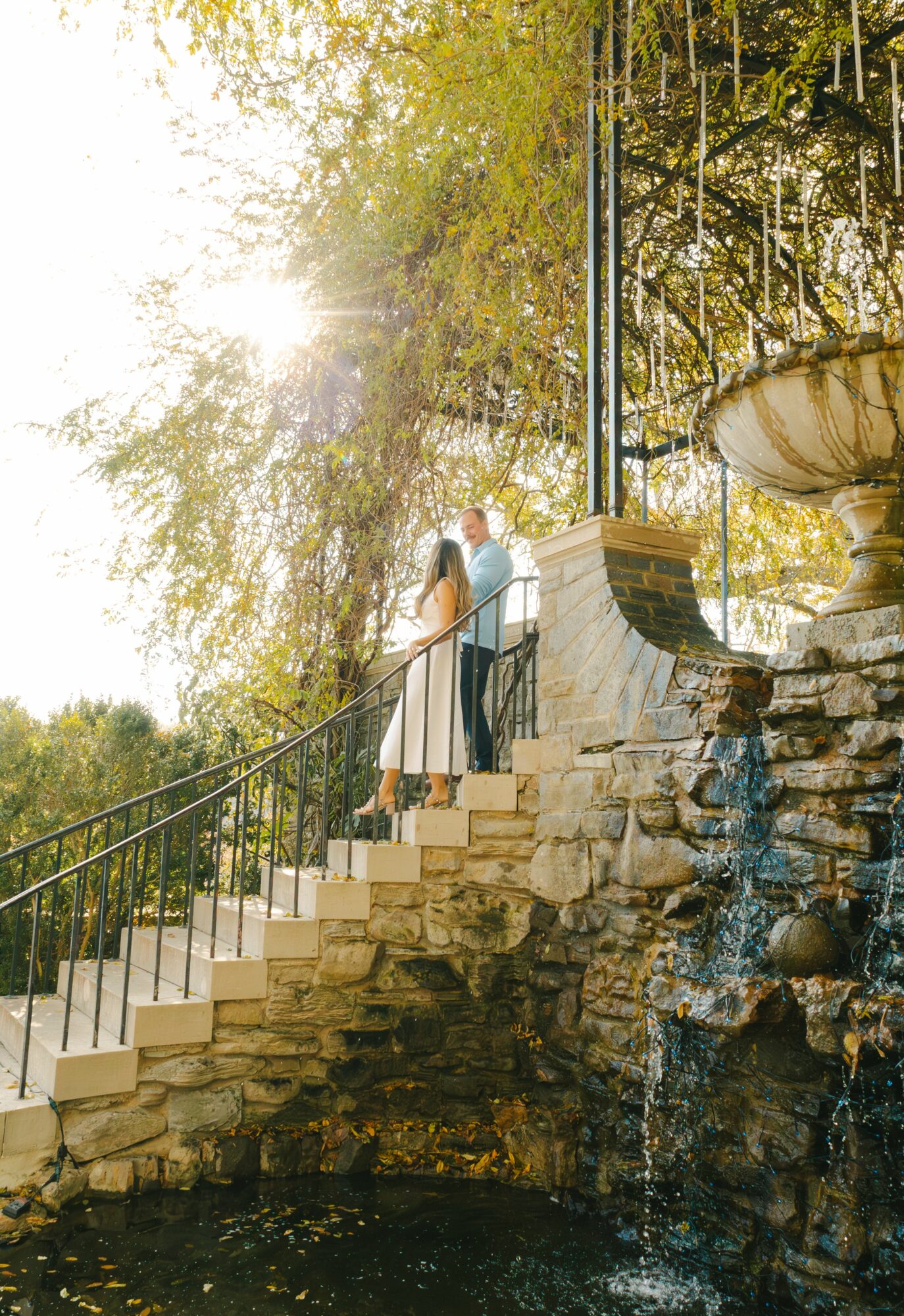 Two people stand on a stone staircase beside a water fountain, with trees and sunlight in the background.