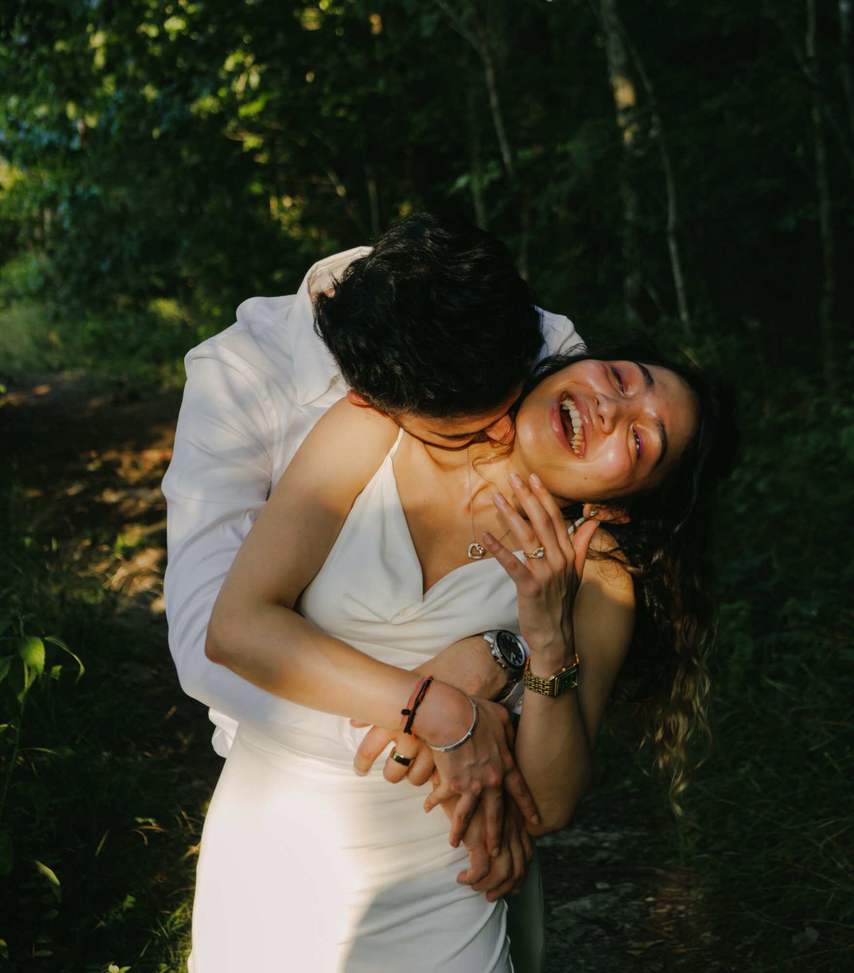 Two people laughing and hugging outdoors in a forested area, sunlight filtering through trees, woman with long dark hair, man with dark hair, both smiling.