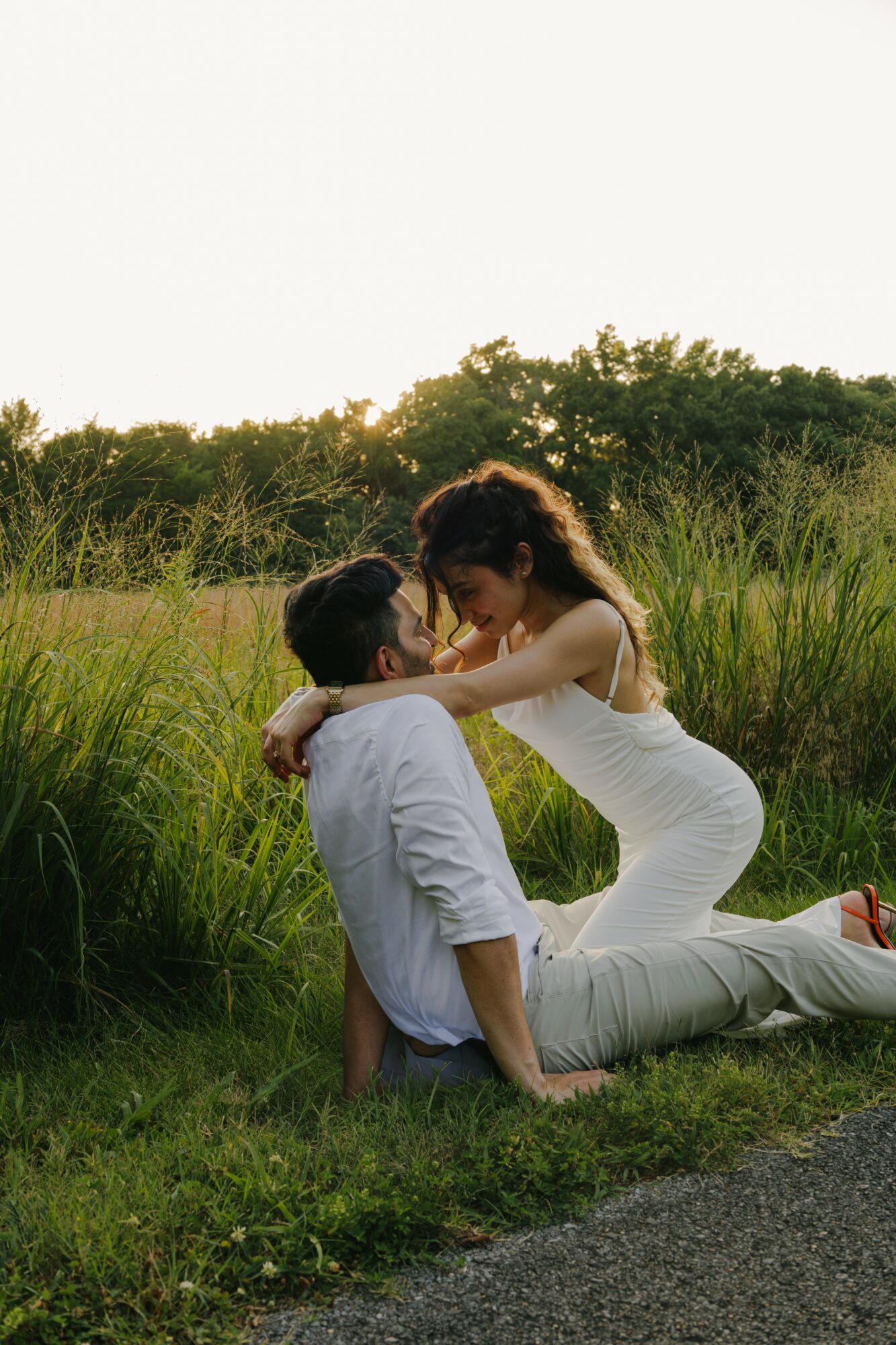 A woman in a white dress and a man in a white shirt and light pants sit on grass, embracing outdoors with greenery in background.