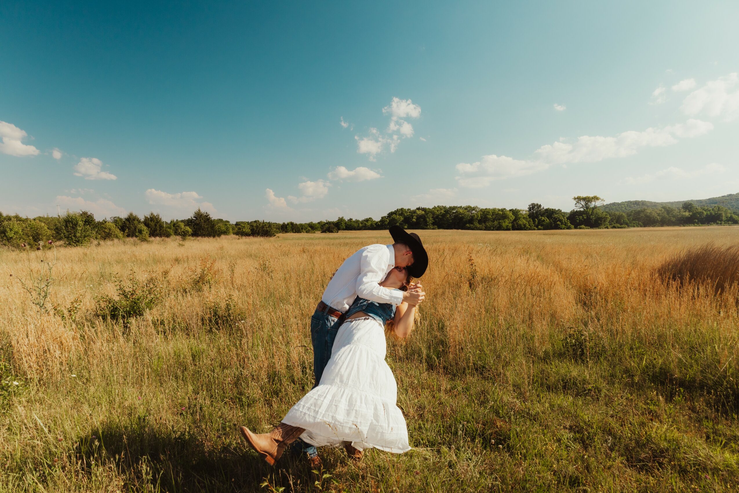 A couple dancing in a grassy field under a blue sky with clouds, holding each other closely.