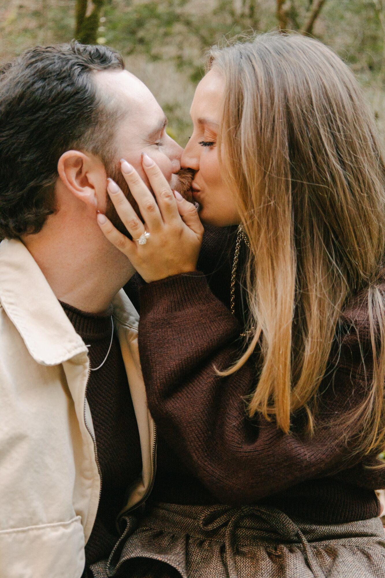 A man and woman are close, touching noses and holding each other's faces outdoors, with trees in the background.