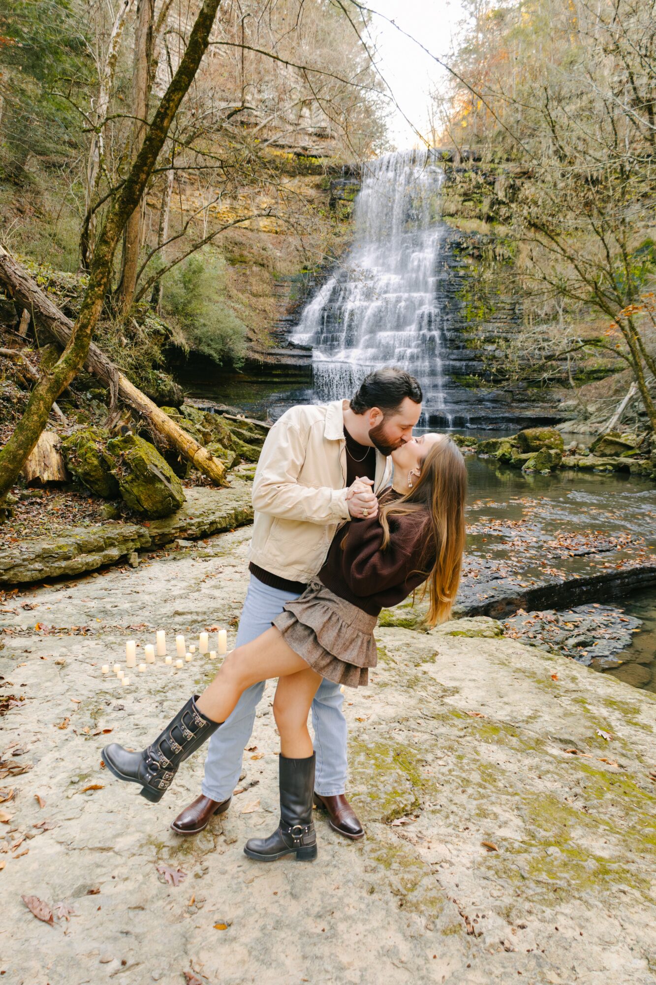 A couple dancing on a rocky path near a waterfall surrounded by trees with autumn leaves.