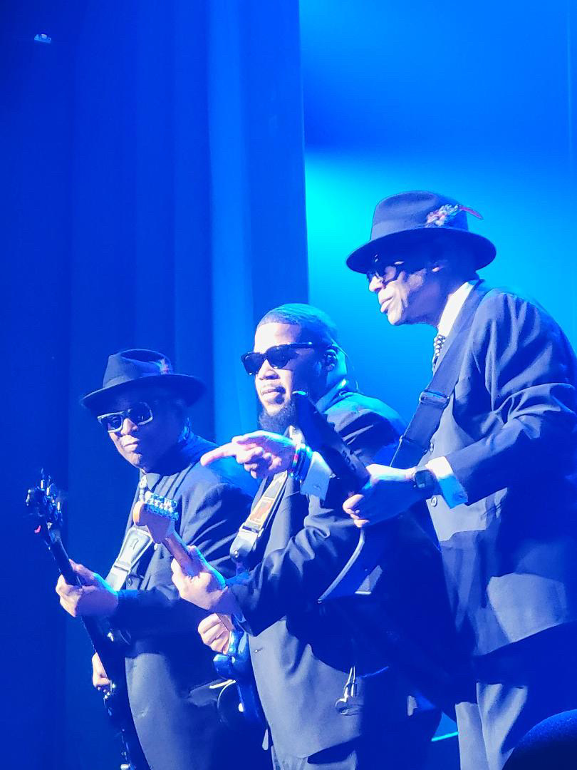 Three musicians in dark suits and hats perform on stage under blue lighting, wearing sunglasses and playing guitars.