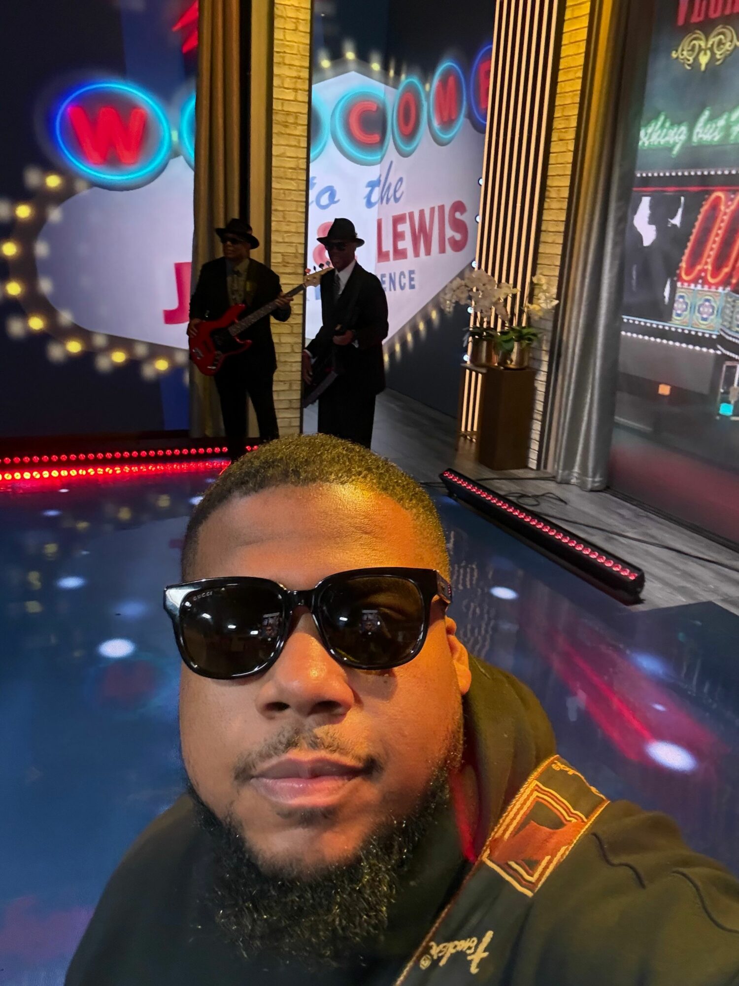 Person with sunglasses and a beard taking a selfie in front of a newsstand with neon signs, nighttime city scene.