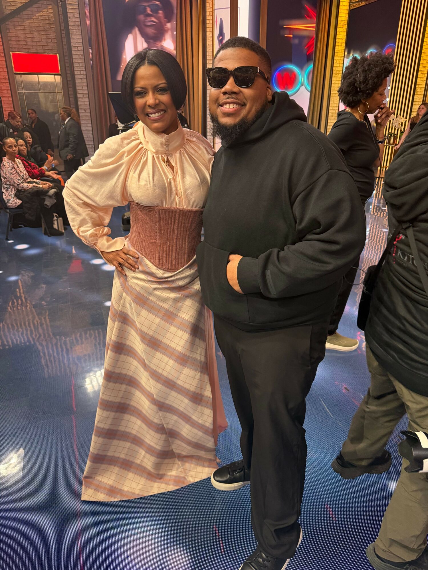 Two people standing in a colorful indoor setting, one wearing sunglasses and a black outfit, the other in a beige top and plaid skirt.