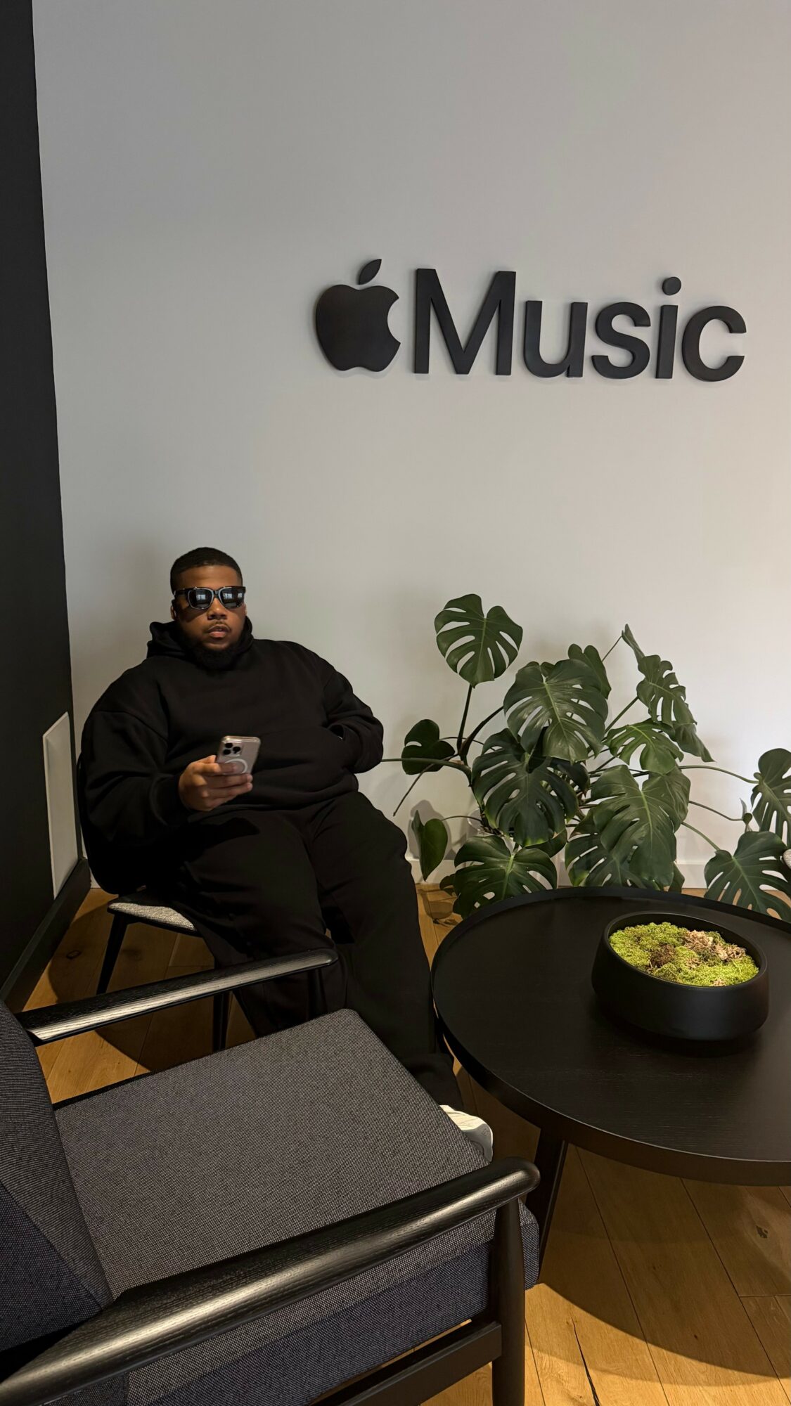 Person sitting on a chair holding a phone, next to a round table with a bowl, in front of a wall with Apple Music logo.