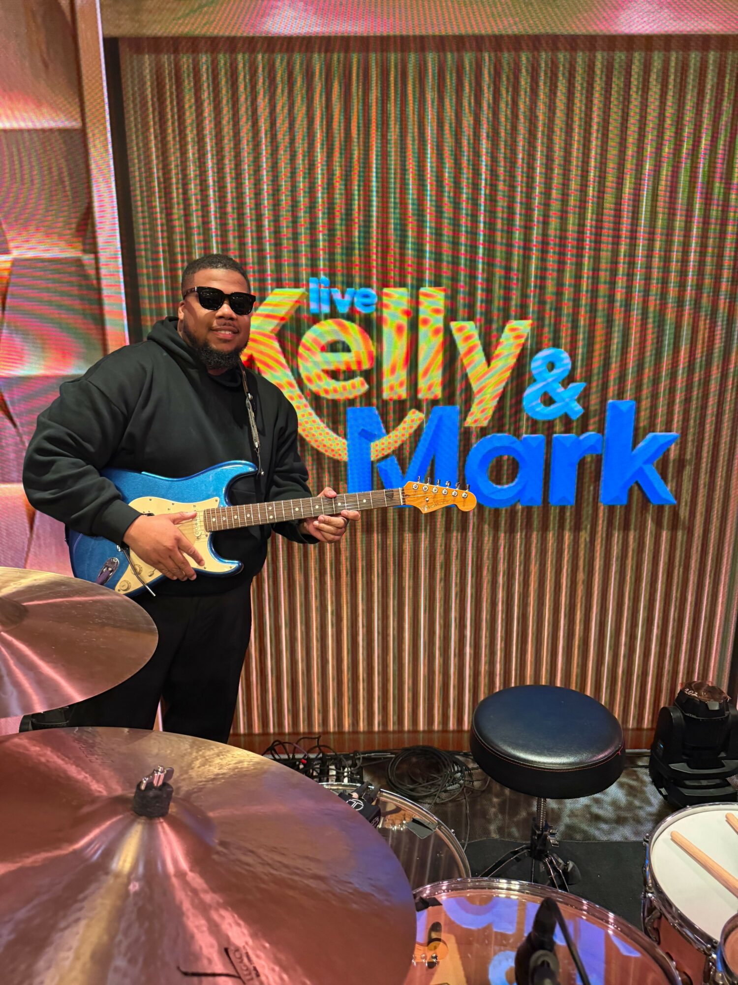 Person playing guitar in front of a colorful striped background, wearing sunglasses and a dark jacket.