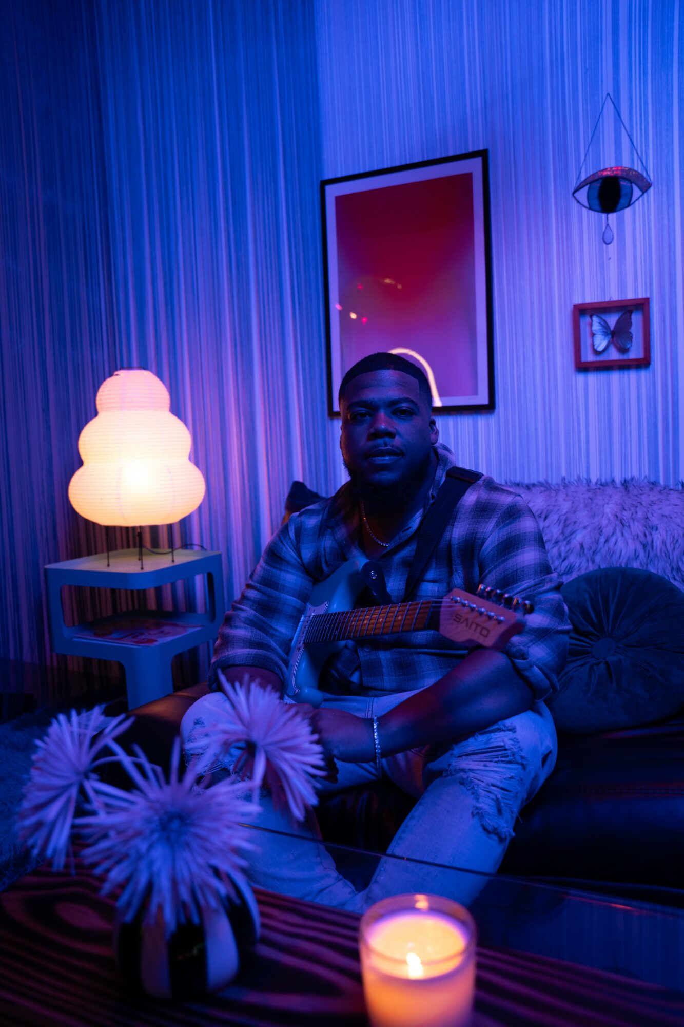 Person sitting on a couch in a room with blue lighting, holding a guitar, surrounded by candles and decorative items.